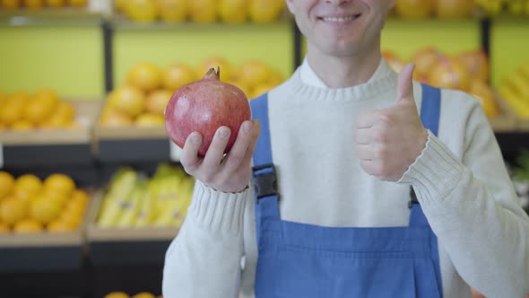 Cheerful Unknown Caucasian Man in Blue Workrobe Holding Pomegranate in Hand and Showing Thumb Up