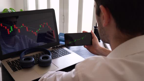 Man Holding Mobile Phone And Looking At Screen Of Laptop Showing Stock Market Bearish Candlestick Mo