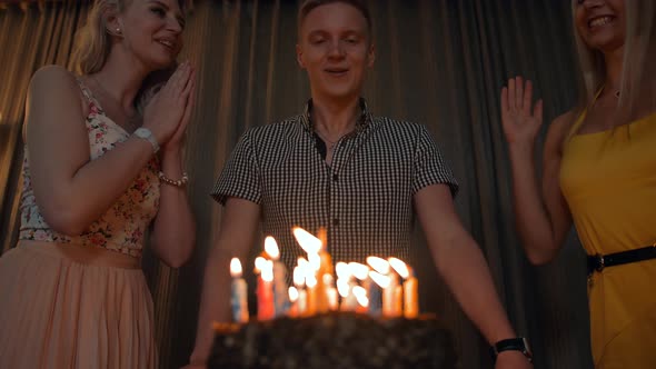 Young Man Blowing Out Much Candles on a Festive Cake with Girls Near.