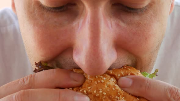 A Young Handsome Man with a Huge Appetite Eats a Big Burger or Cheeseburger on the Terrace of a