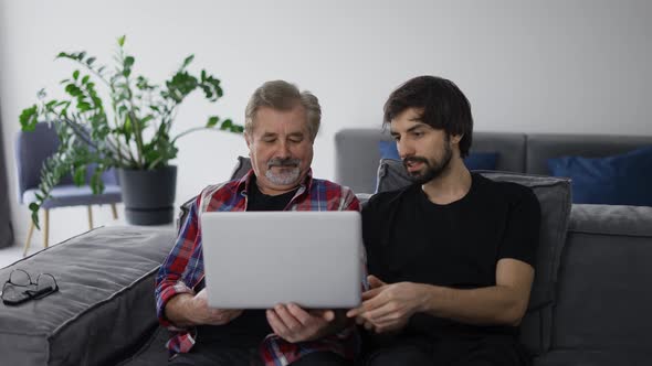 Young Man Teaching Old Dad Using Internet Online with Computer on Couch in Living Room