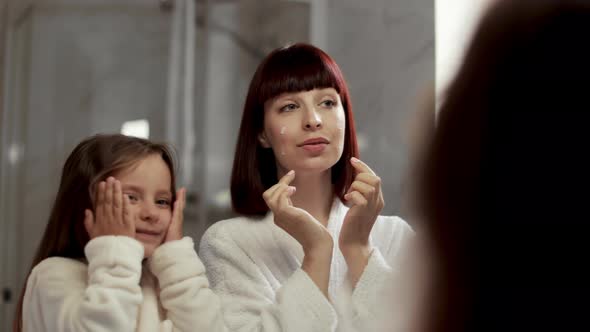 Content Young Woman and Little Daughter in White Bathrobes Applying Cream on Cheeks