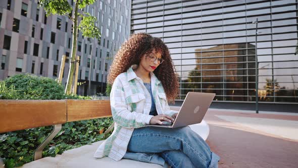 Curly African American Girl with Afro Hairstyle Sitting Outdoors on Street in the City in Business