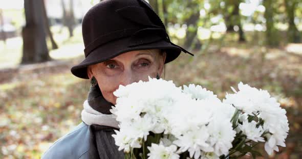 Happy Elderly Woman in Coat and Hat Sniffing a Bouquet of Flowers and Smiling