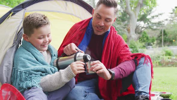 Happy caucasian father with son sitting in tent and drinking tea in garden