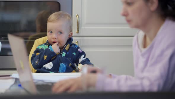 Mother Works on Laptop While Her Toddler Sitting in High Chair Next to Mother