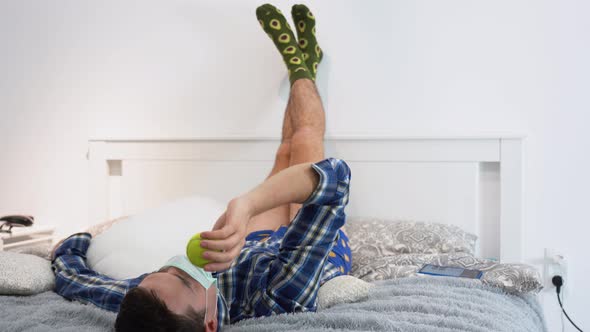 Idle man wear medical face mask, lies on bed, throws up, catches yellow tennis ball. Quarantine