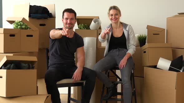 A Moving Couple Sits on Chairs in an Empty Apartment, Smiles and Points at the Camera