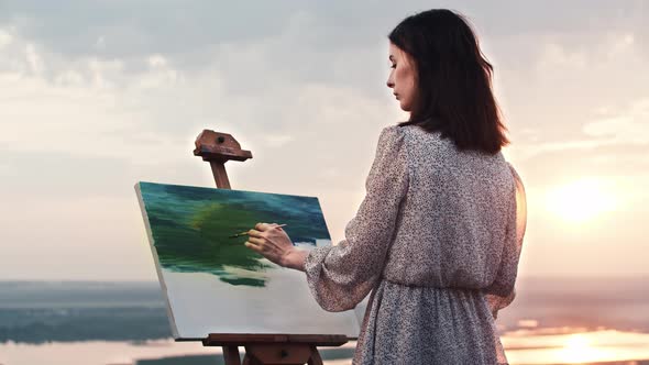 A Young Woman Artist in Light Dress Standing at Sunset and Drawing a Painting of Nature Landscape