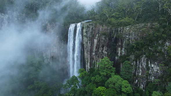 Unique drone view through suspended mist revealing a majestic waterfall spilling down a lush rainfor