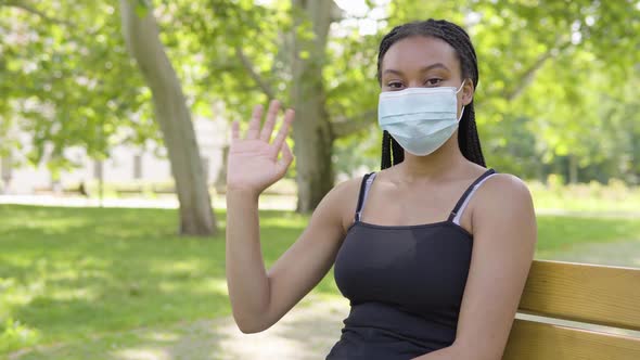 A Young Black Woman in a Face Mask Waves at the Camera and Sits on a Bench in a Park on a Sunny Day