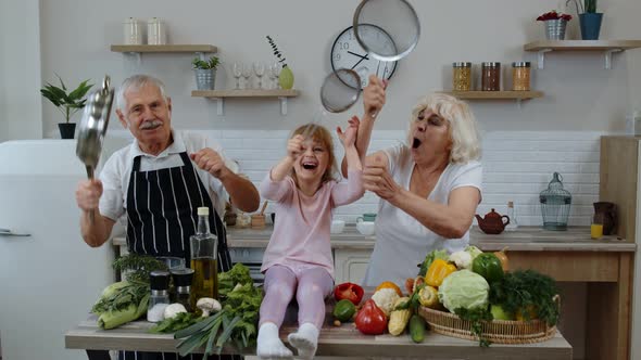 Happy Vegan Senior Couple Dancing with Granddaughter Child While Cooking Vegetables in Kitchen