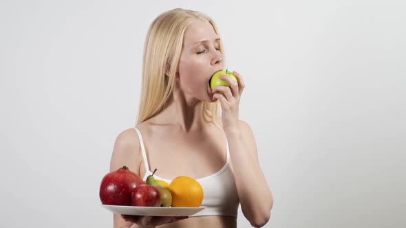 Young Attractive Woman with a Plate of Fruit in Her Hands Eats an Apple on a White Background