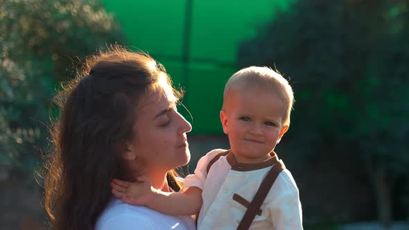 Young Mother Holds Little Son in Arms Standing in Backyard