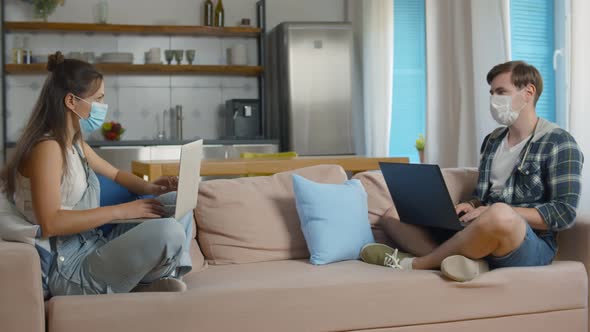 Couple in Medical Masks Using Laptops on Couch Staying at Home