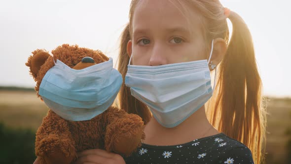 Sad Little Girl and Teddy Bear with Surgical Mask at Sunset. A Child Holding Her Teddy Bear During