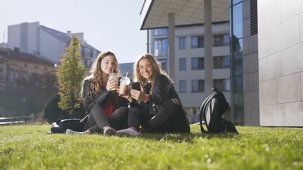 Young Caucasian Women Drinking Fresh Juice while Sitting on the Green Grass Using Mobile Phone