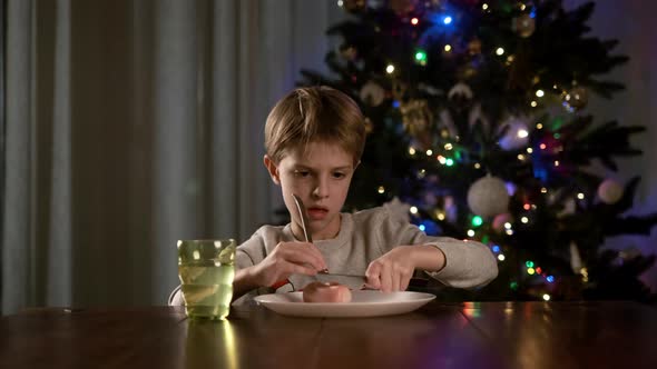 New Year, Funny Boy Sits At Table And Hardly Cuts Sausage On Plate, In The Background Is Christmas