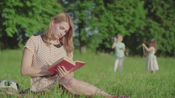 Positive Beautiful Mother Reading Book on Green Meadow and Looking Back at Children Playing at the