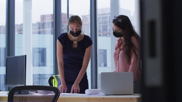 Two diverse female colleague wearing face masks standing at table looking at blueprints and talking