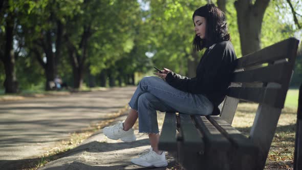 Side View Caucasian Teenage Girl Sitting on Bench in Summer Park Surfing Social Media on Smartphone