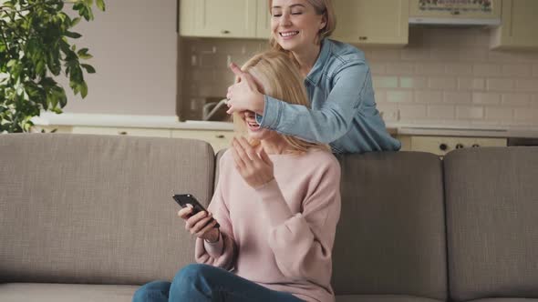 Smiling Young Adult Daughter Makes an Unexpected Surprise to Her Middle Aged Attractive Mom Gives