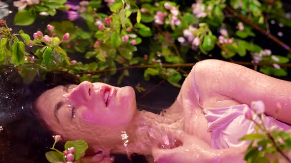 Fascinating Excited Young Woman is Lying in Water with Blooming Tree Branches in Picturesque Lake