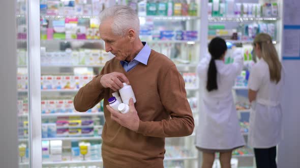 Senior Man Collecting Bottles with Pills on Shelf in Drugstore Leaving with Blurred Pharmacists