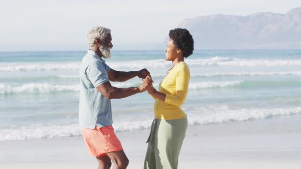 Happy african american couple dancing and holding hands on sunny beach