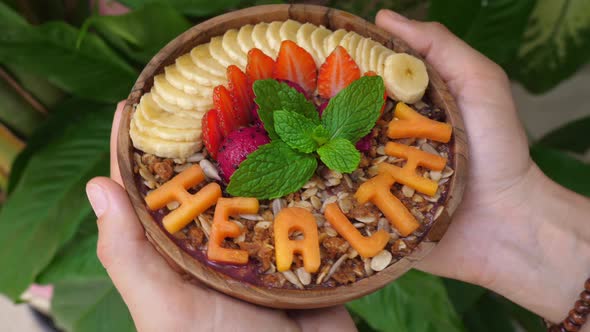 Hands Holding a Coconut Smoothie Bowl Topped with Granola and Word "Healthy" Carved Out of Fruits
