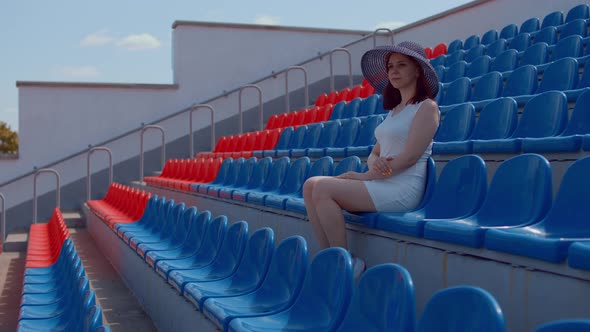 Young Woman in in White Dress and Elegant Hat Looks Away Sitting on Stadium Bleachers Alone