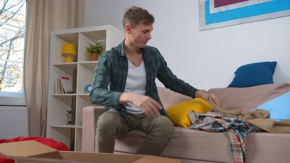 Young Handsome Guy Sitting on Couch and Packing Donation Box with Old Clothes