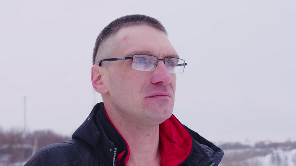 An Adult Man with Glasses Looks Thoughtfully Into the Distance Standing on the Street in Winter in