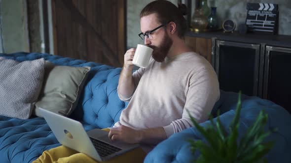 Man Using Laptop Sitting on Couch at Home Spbd. Thinking Hipster Guy with Device Looking at Screen