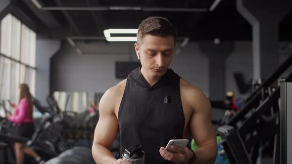 Young Caucasian Man of Athletic Physique Walks Through the Gym with a Smartphone in His Hand