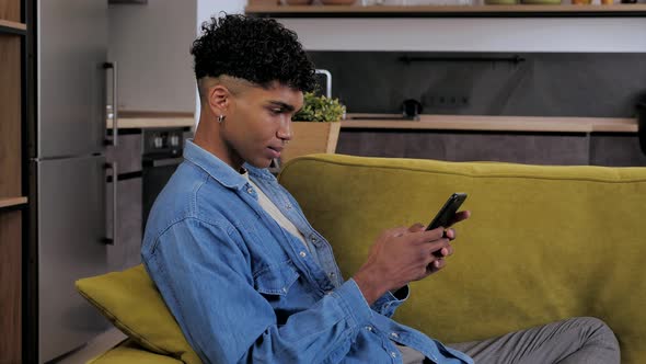 Young Stylish Millennial Afro American Boy in White TShirt Using His Smartphone for Chatting Typing