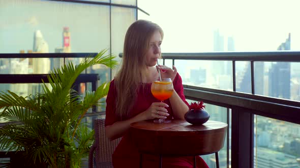 Elegant Young Woman with Cocktail Rests at Luxury Rooftop Restaurant