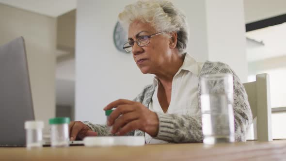 African american senior woman holding empty medication container while using laptop at home