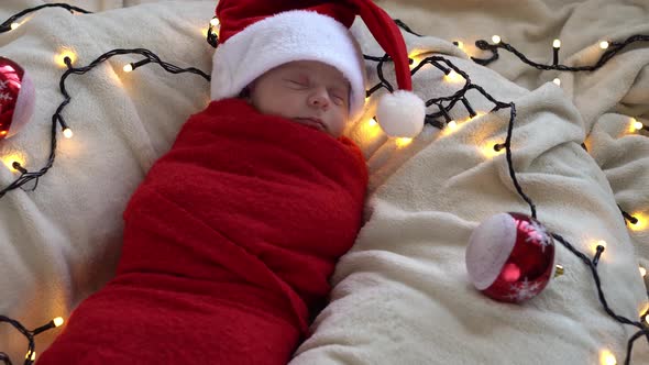 Top View Portrait First Days Of Life Newborn Cute Funny Sleeping Baby In Santa Hat Wrapped In Red