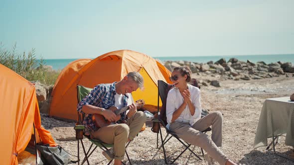 Handsome Man Checkered Shirt Is Sitting in Camp Chair with Ukulele and Sings Funny Song for His