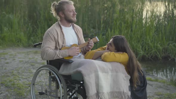 Camera Approaches To Smiling Disabled Man on Wheelchair Playing Uklele for Beautiful Young Woman