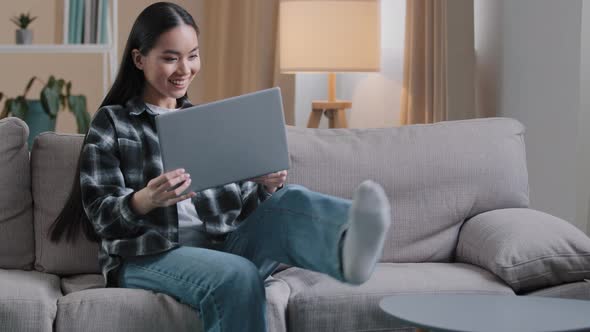 Carefree Asian Girl Enters Living Room Sit Down on Comfortable Sofa Takes Laptop with Smile and