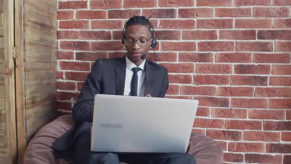 Black Man in Headset Glasses and Strict Suit Speaks Via Video Link Through a Laptop Sitting on Bag