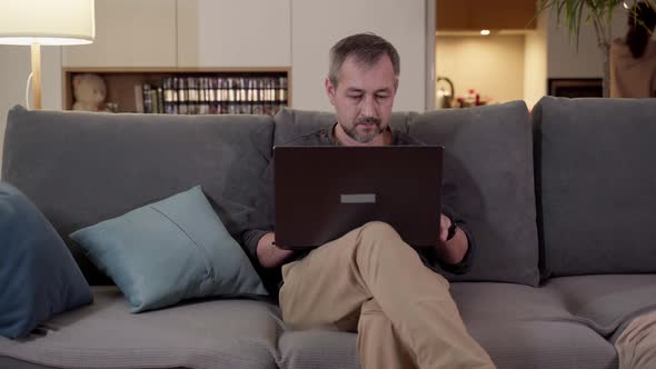 In the Evening a Man with a Laptop Sits on the Couch