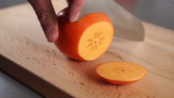 The hand of a young man slices with a knife a ripe orange persimmon on a wooden cutting board