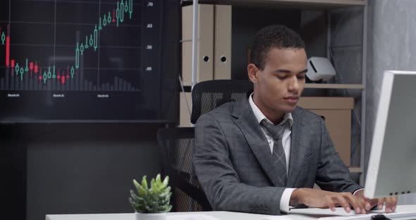 Young African Man in a Modern Office, Working at the Computer, Screens with Financial Infographics