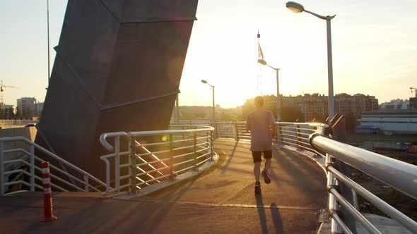 a Guy in a Tshirt and Shorts is Runing Across the Bridge at Dawn or Sunset
