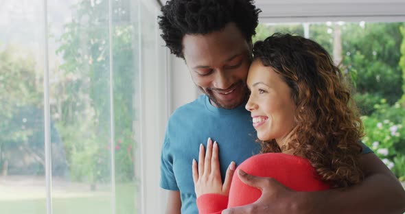 Happy biracial couple embracing, standing at window and looking into distance