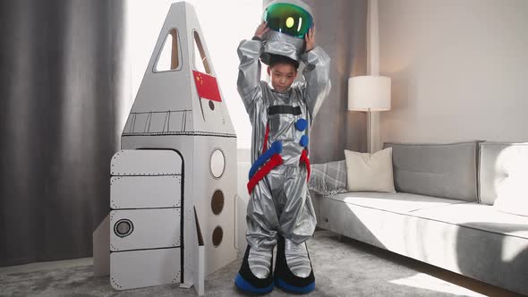 An Asian Boy in an Astronaut Costume Stands Near a Cardboard Model of a Spaceship and Puts Off