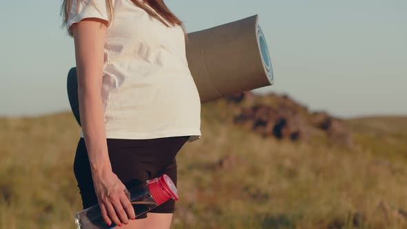 Pregnant White Woman Stands Outdoor and Holds Fitness Mat and Bottle of Water in Her Hands Closeup